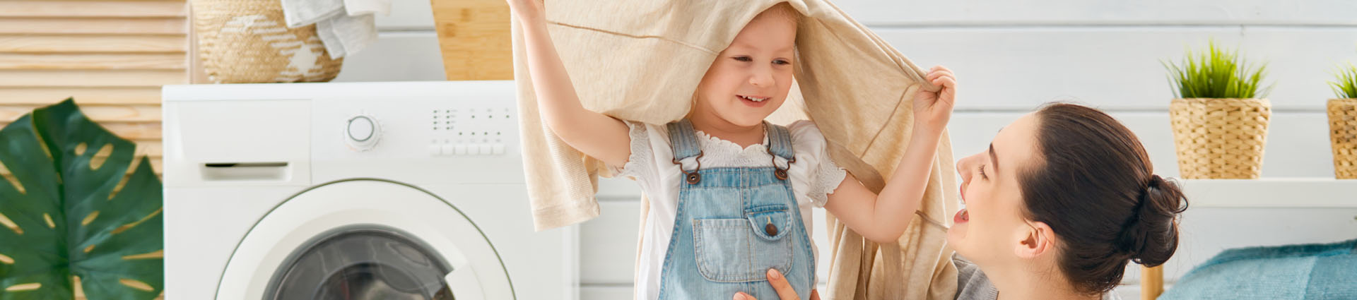 smiling child peaking from underneath a towel draped over their head with laughing woman sitting in laundry room